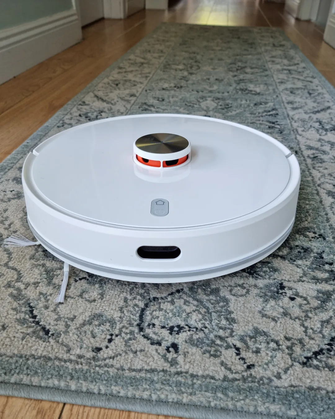 Robot aspirador redondo y blanco sobre una alfombra estampada en tonos gris y azul, en un pasillo con suelo de madera
