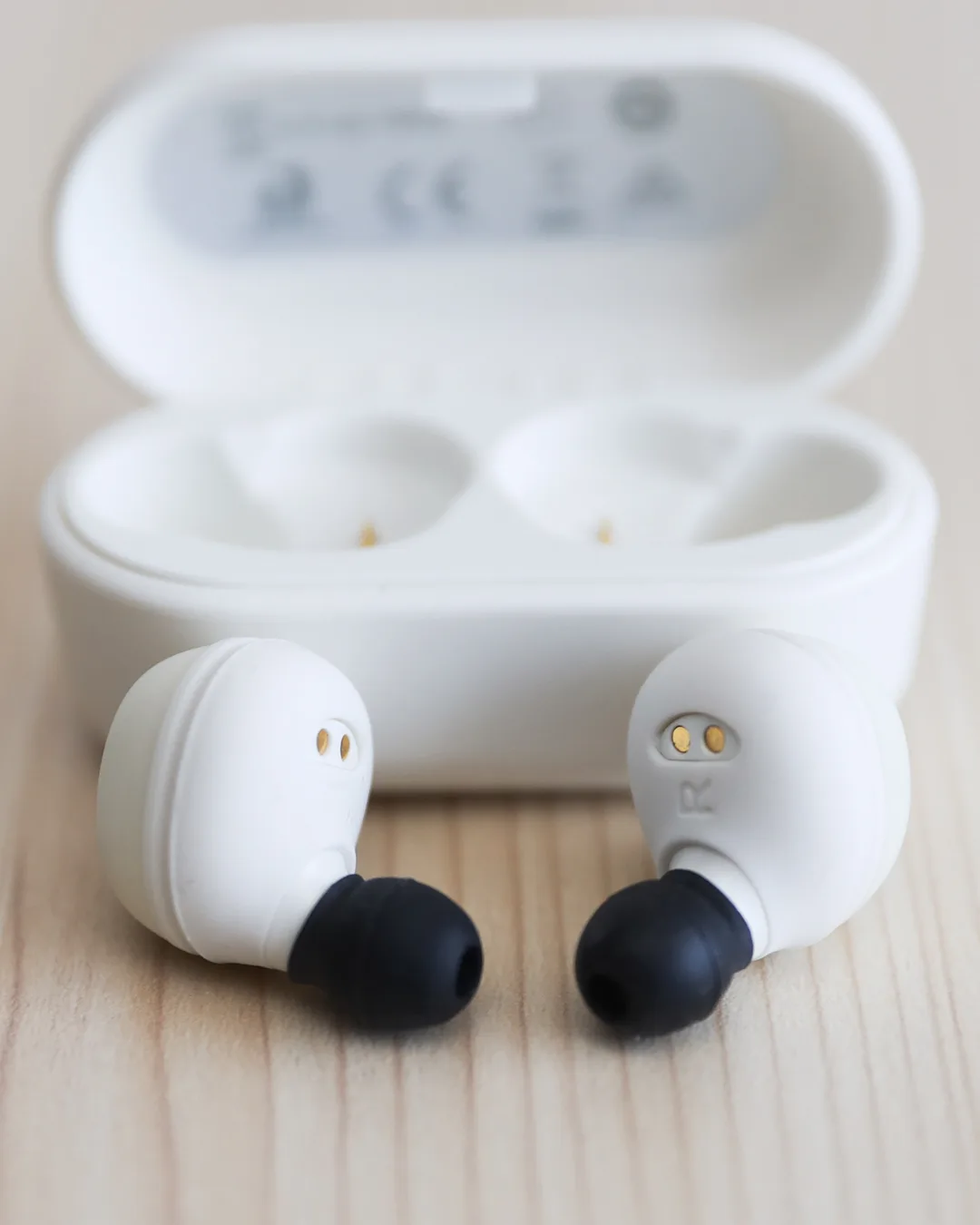 Dos auriculares inalámbricos blancos de botón, con almohadillas negras, delante de su estuche de carga abierto sobre una mesa de madera clara